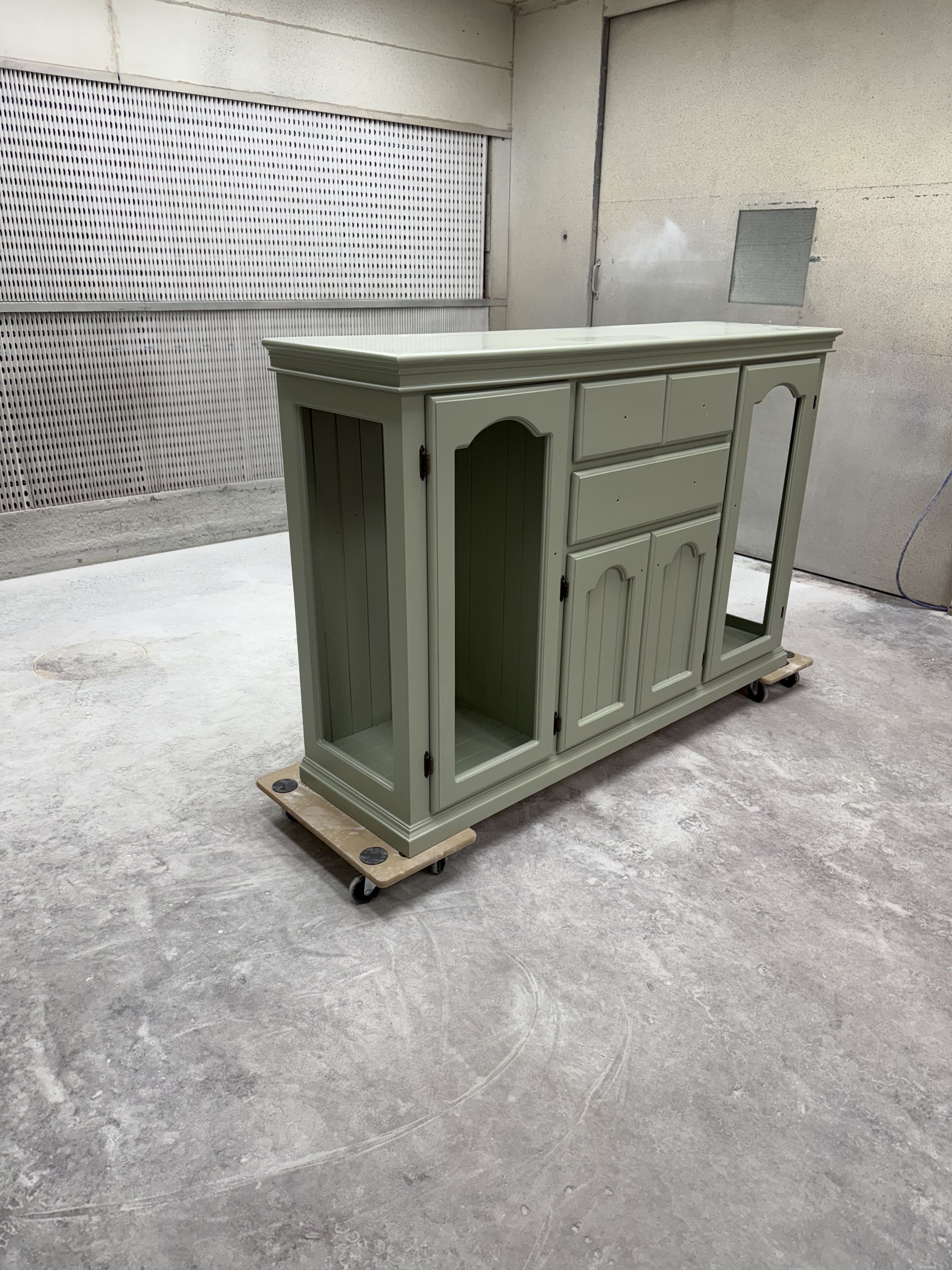 A painted green cabinet on a wheeled platform inside a spray booth. The cabinet has doors, drawers, and glass panels.