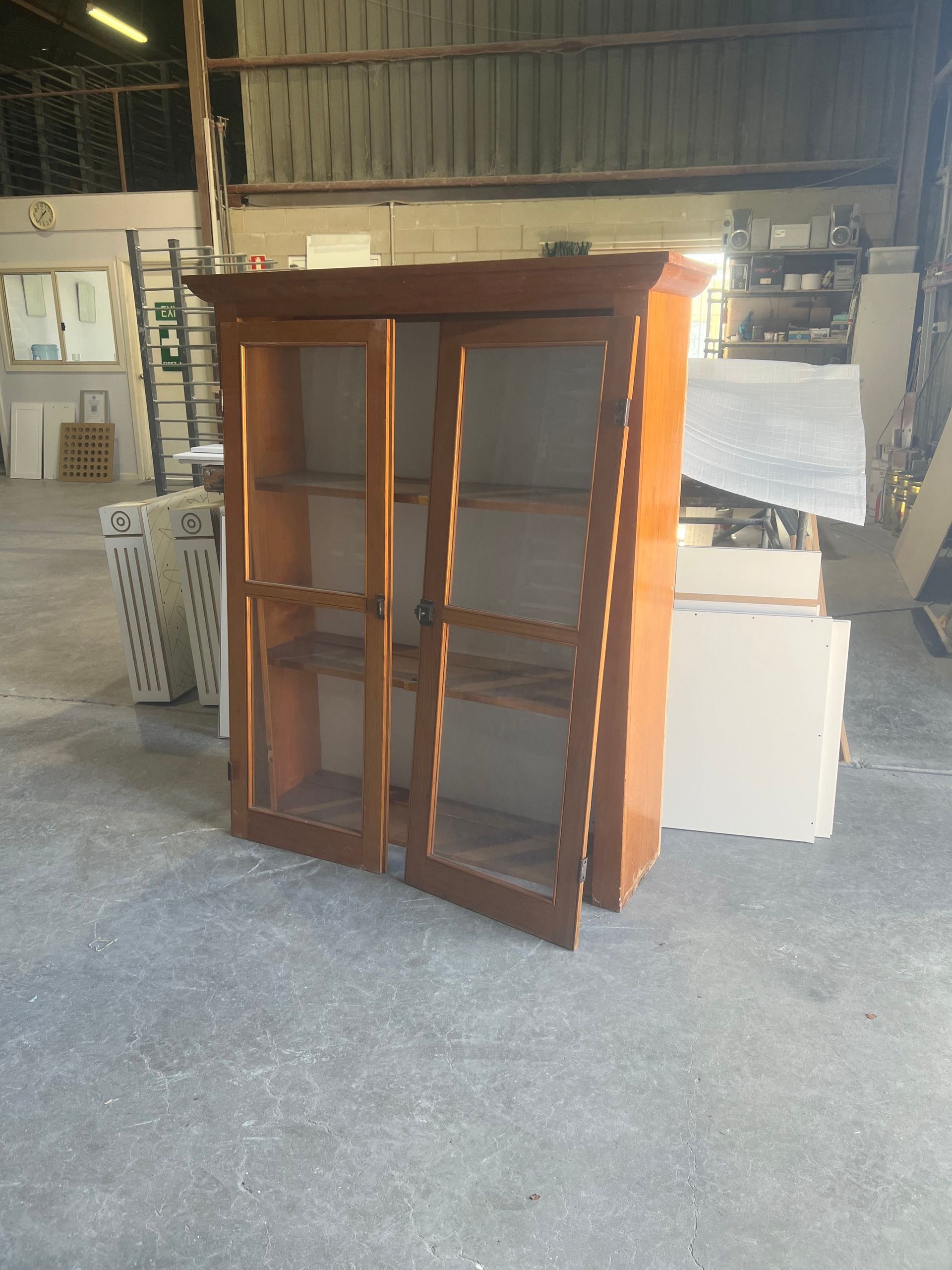 Wooden cabinet with glass doors and shelves, in a cluttered warehouse.