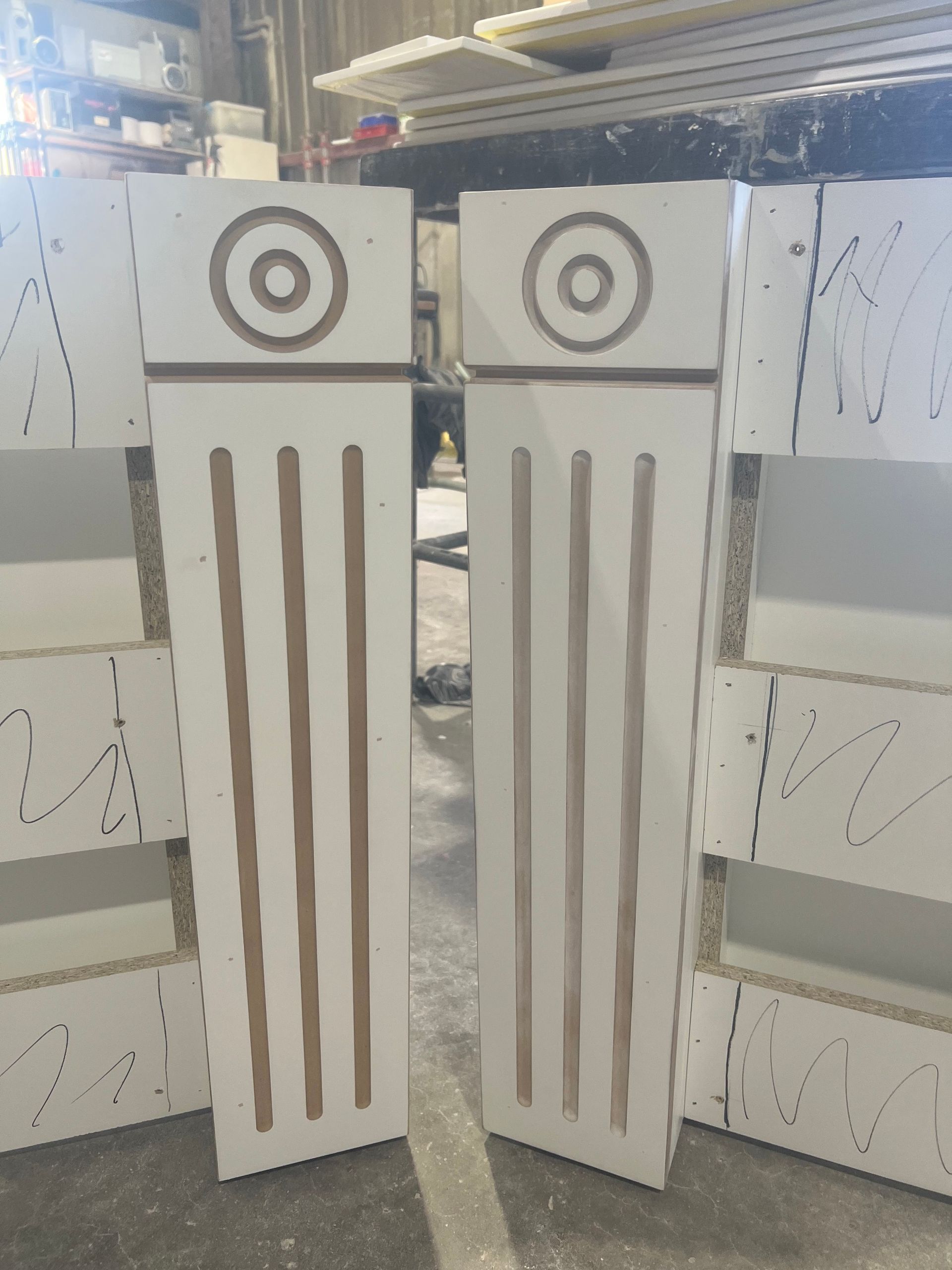Two white wooden pillars with brown grooved lines and circular designs at the top, in a workshop setting.