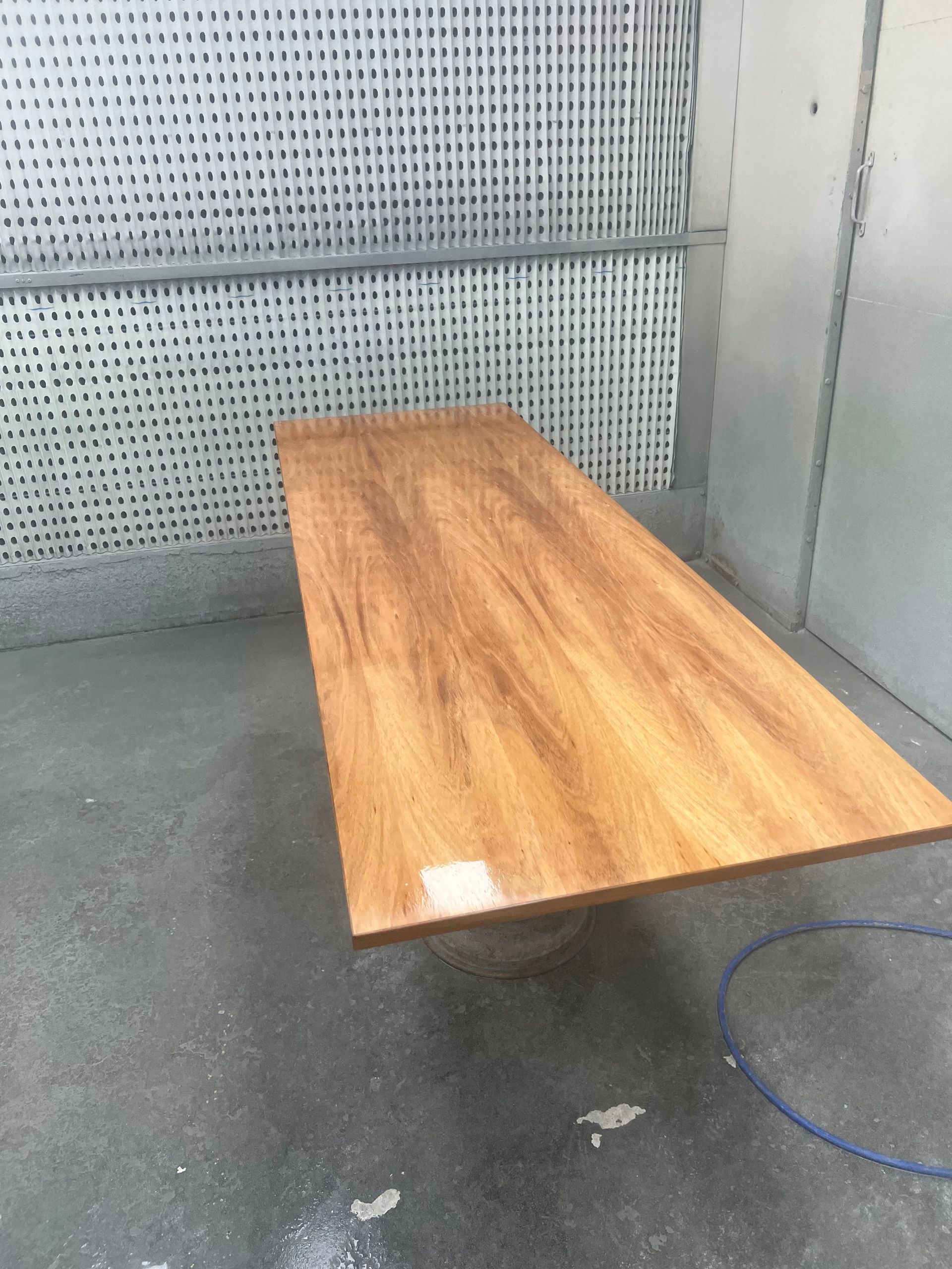A wooden table with a glossy finish is in a spray booth. The table is positioned on a circular base with the walls behind it.