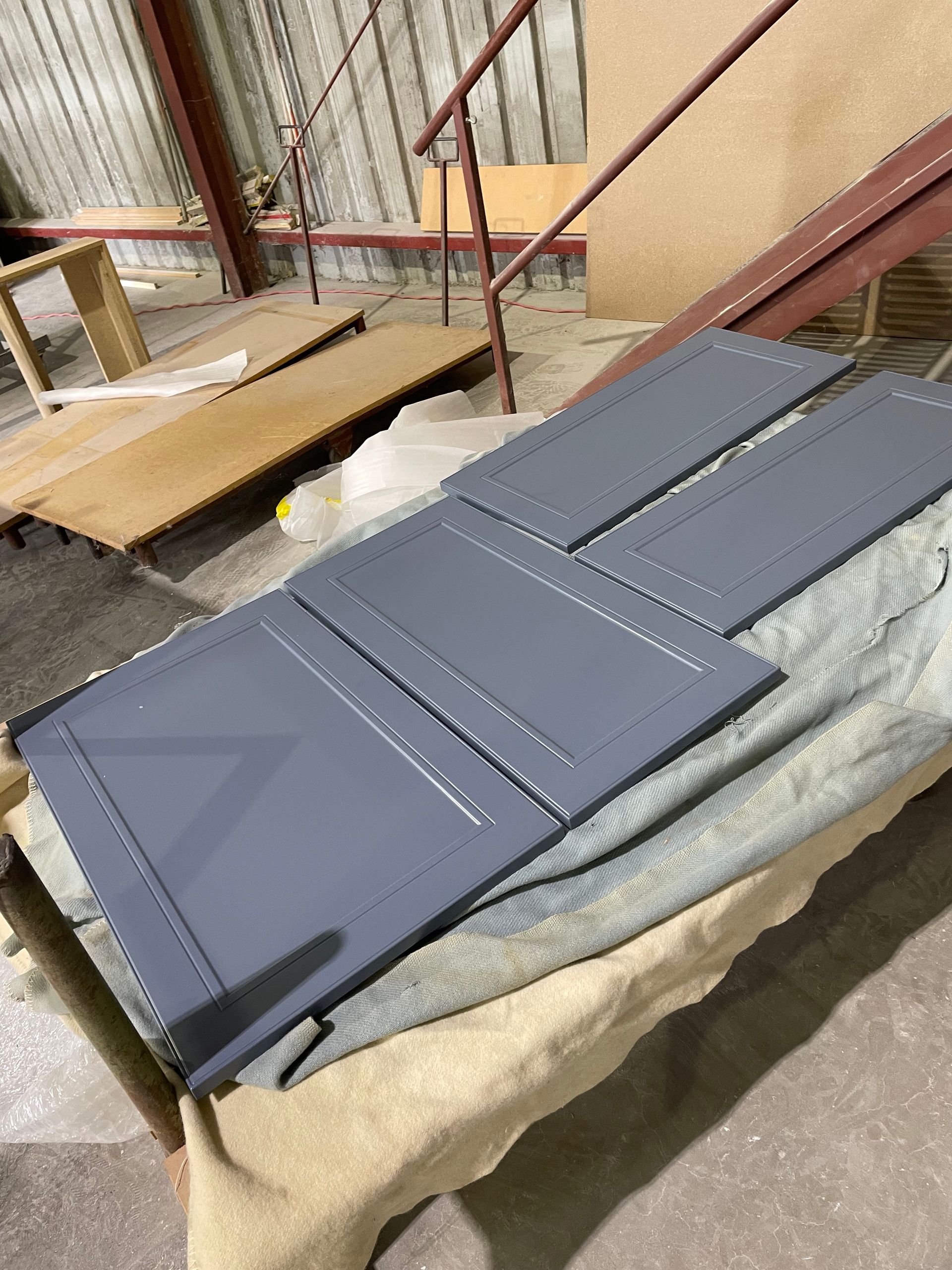 Four gray cabinet doors lying on a fabric surface in a workshop setting, with wooden boards and a ladder in the background.