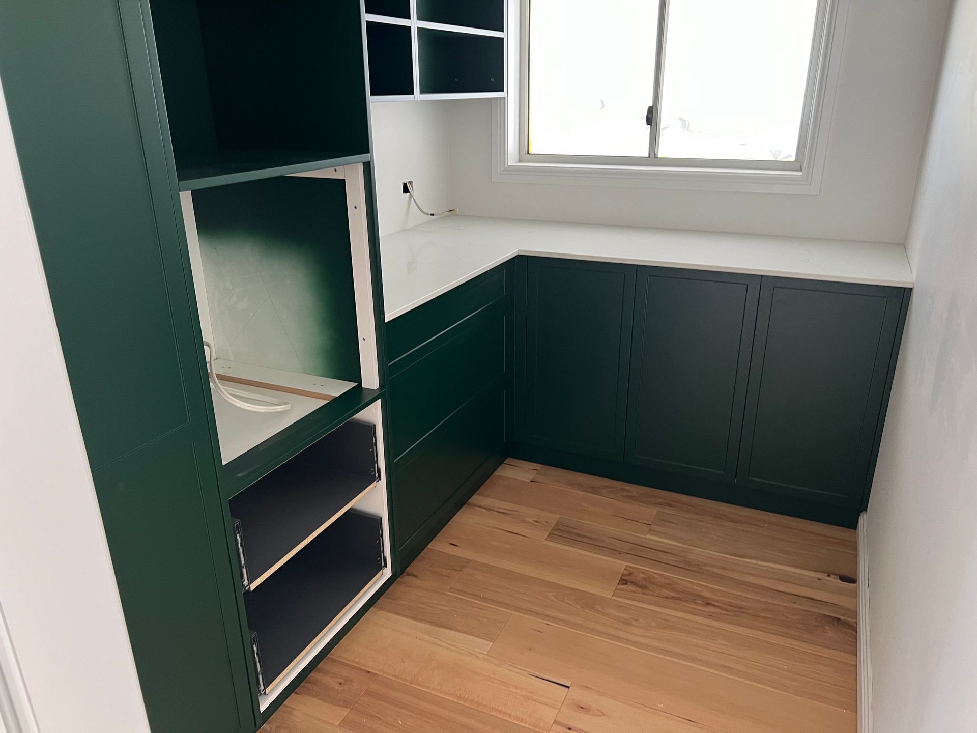Interior of a pantry with dark green cabinets, a white countertop, and wooden floors. A small window is above the countertop.