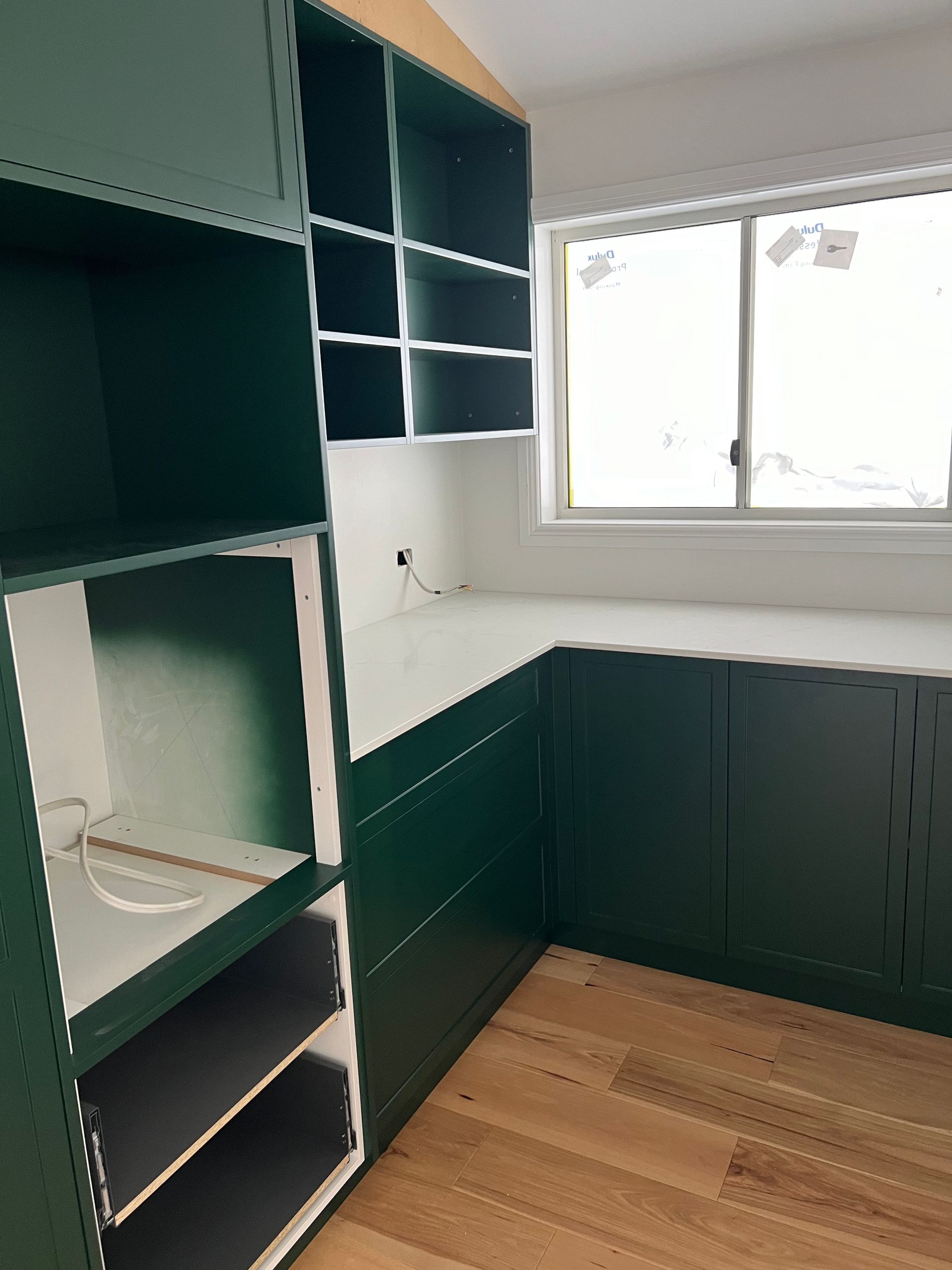 Green built-in cabinets with open shelves and a countertop, next to a window in a room with light wood flooring.