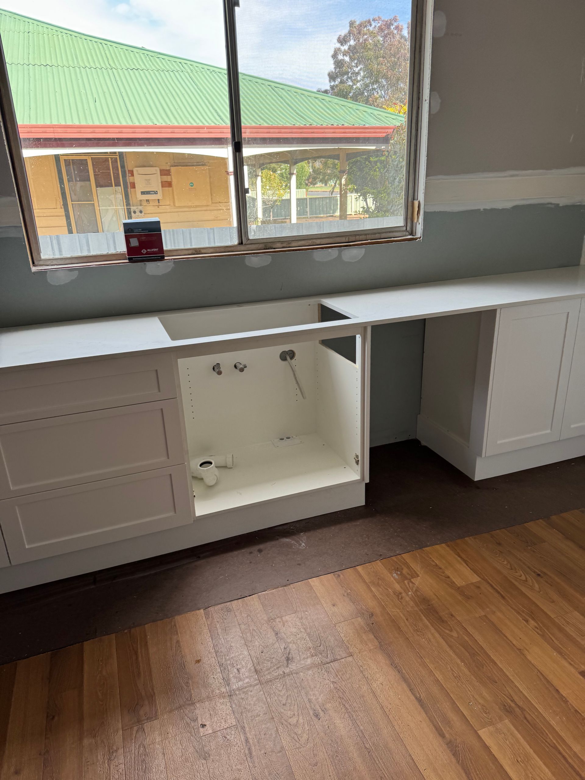 White kitchen cabinets with an open sink area beneath a window, in front of a wooden floor. The window overlooks a building with a green roof.