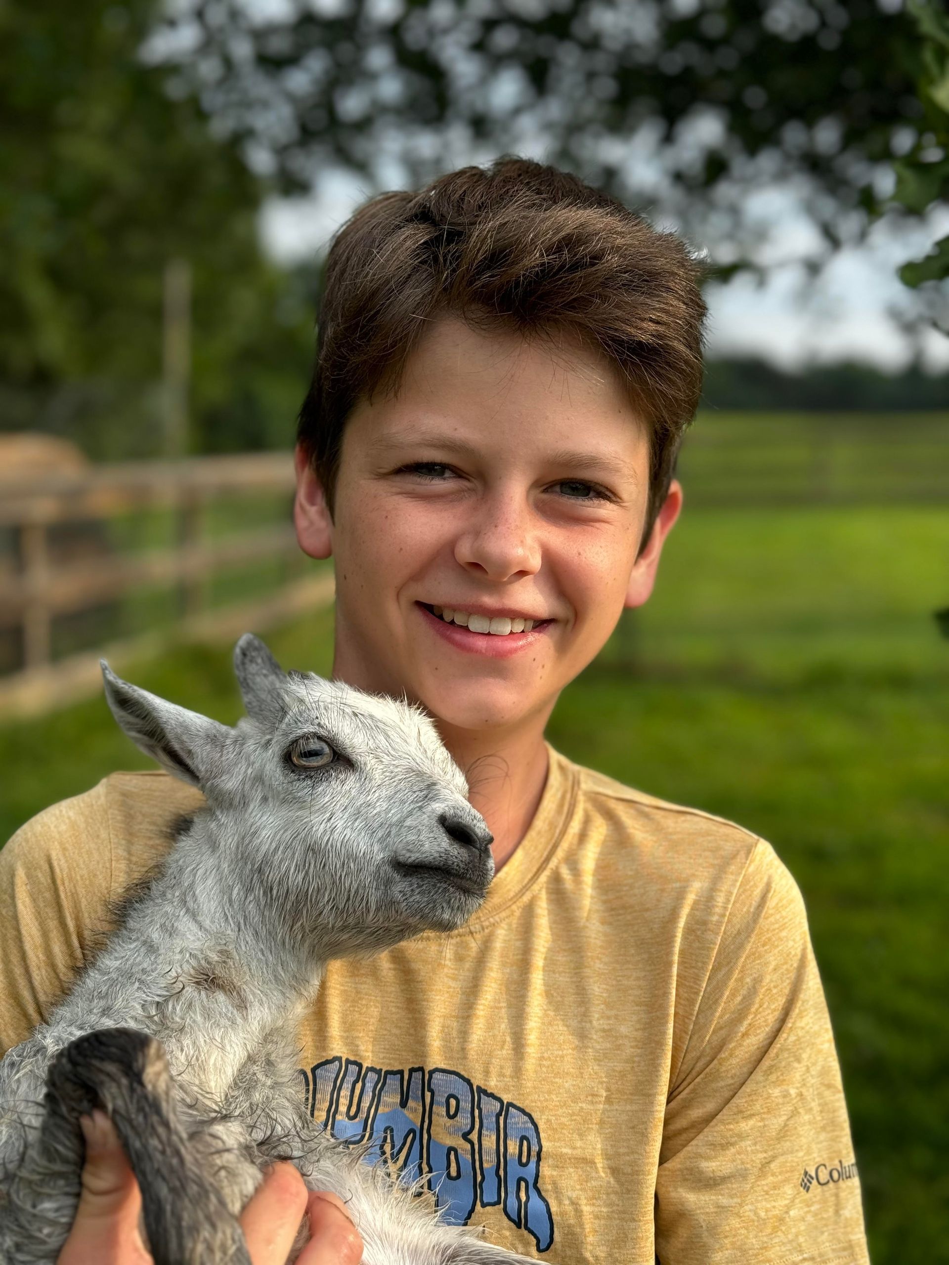 Boy holding a baby lamb, both smiling. Outdoors, field, fence.