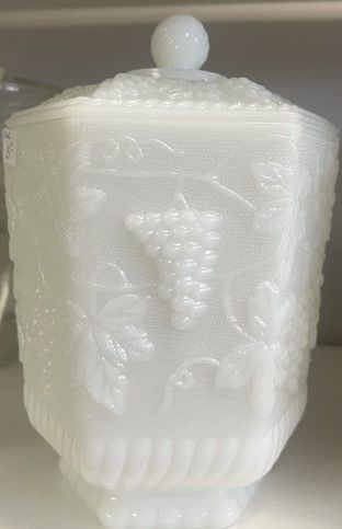 A white glass container with a grape design on it is on a shelf.