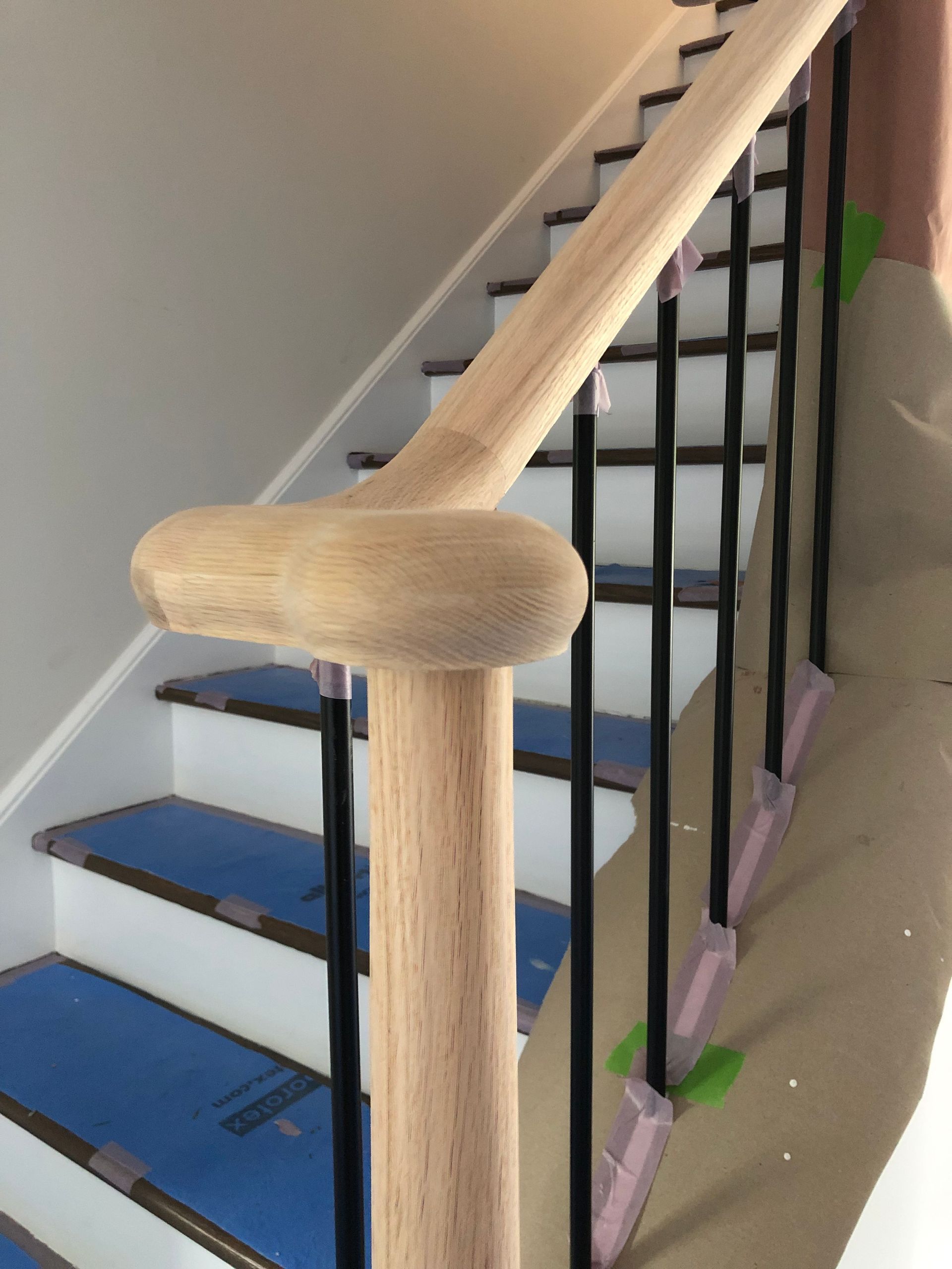Close-up of wooden handrail and black metal balusters on a staircase under renovation, with protective sheets and tape visible.
