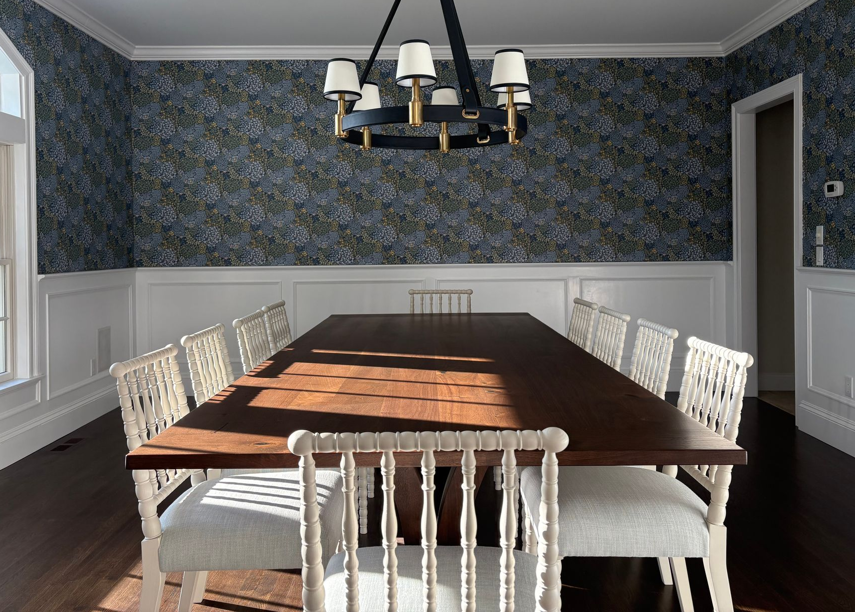 Elegant dining room with floral wallpaper, white wainscoting, and wooden table surrounded by white chairs under a black and gold chandelier.