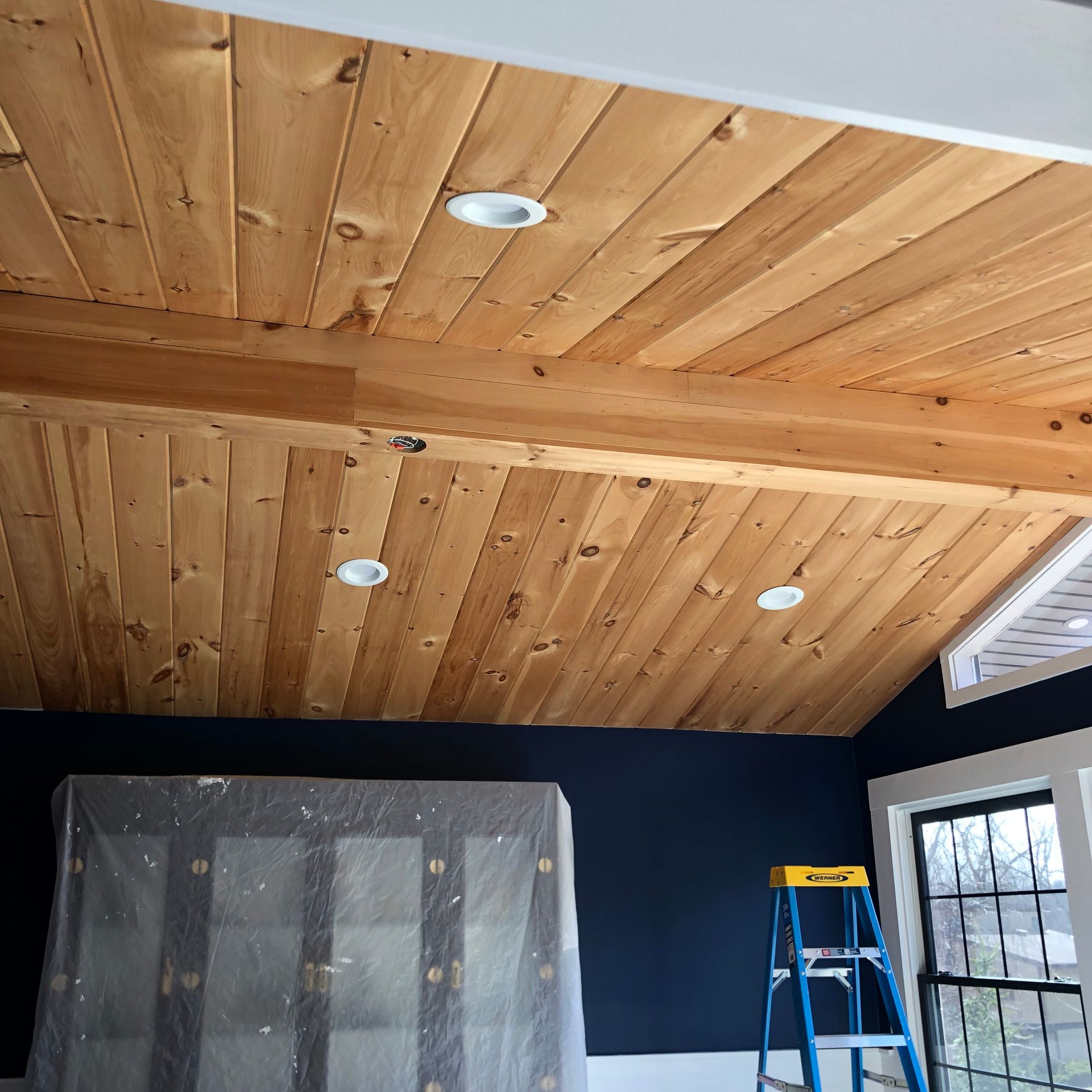 Room with vaulted wood ceiling, recessed lighting, and dark blue walls, featuring a step ladder and covered furniture during renovation.