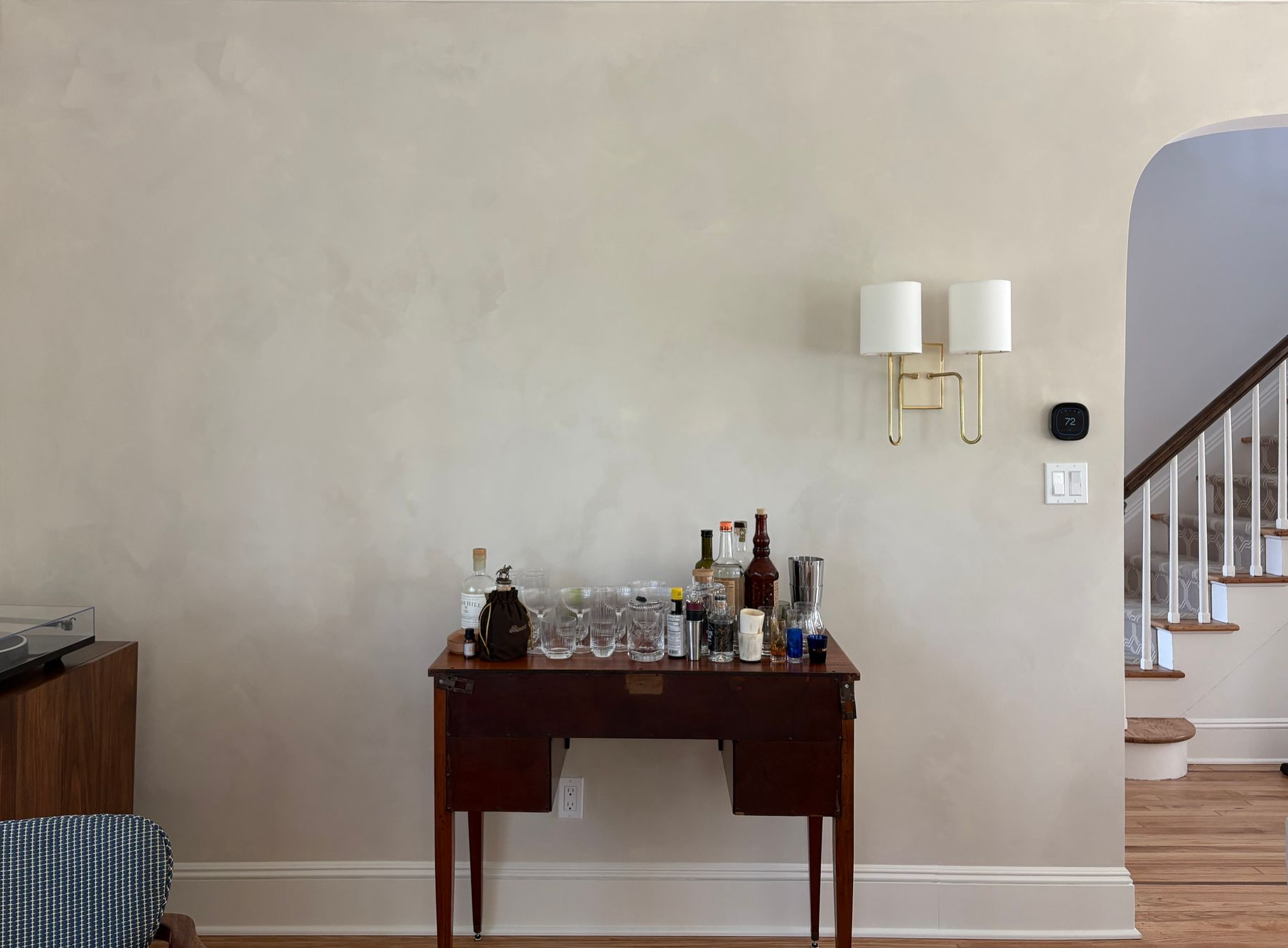 Stylish home bar with liquor bottles and cocktail accessories on a wooden table beneath a modern light fixture, set against a beige wall near a staircase.