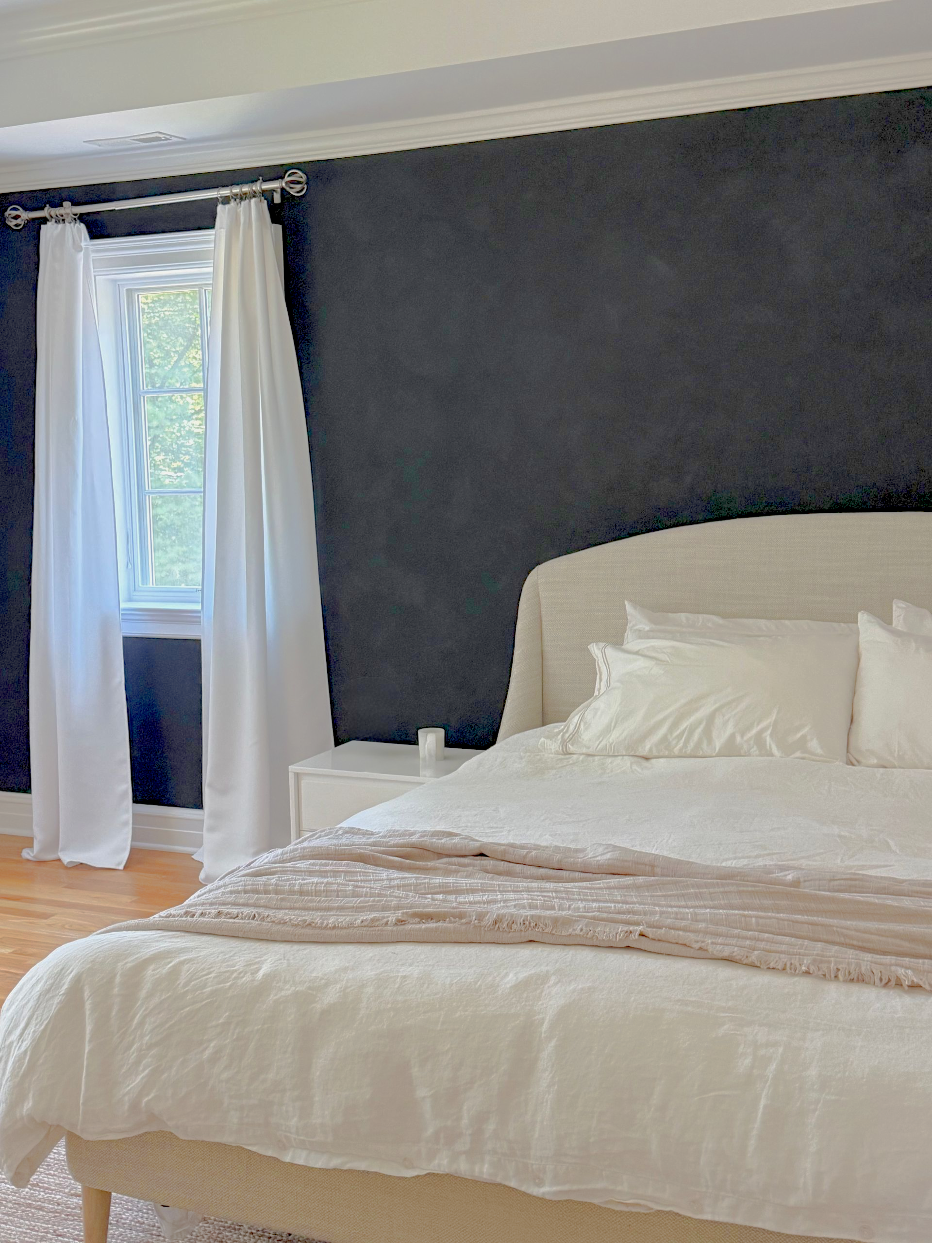 Modern bedroom with white linens, beige throw, and dark charcoal accent wall, illuminated by natural light through window with white curtains.