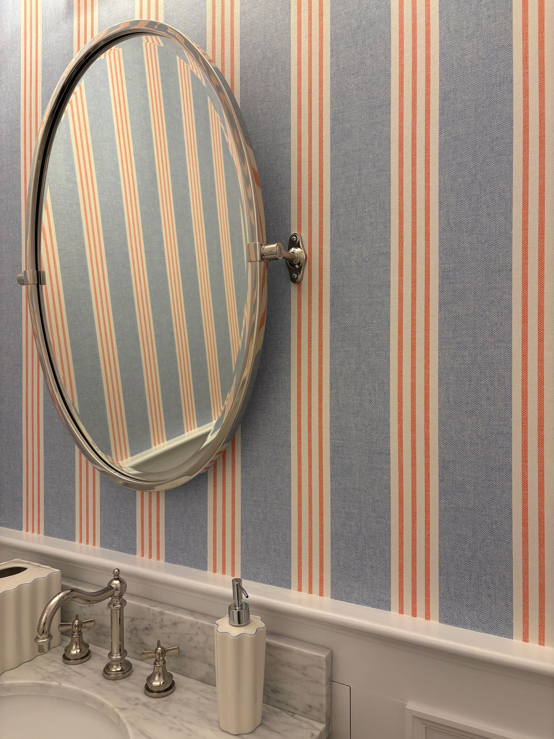 Vertical striped wallpaper behind round mirror and marble sink.
