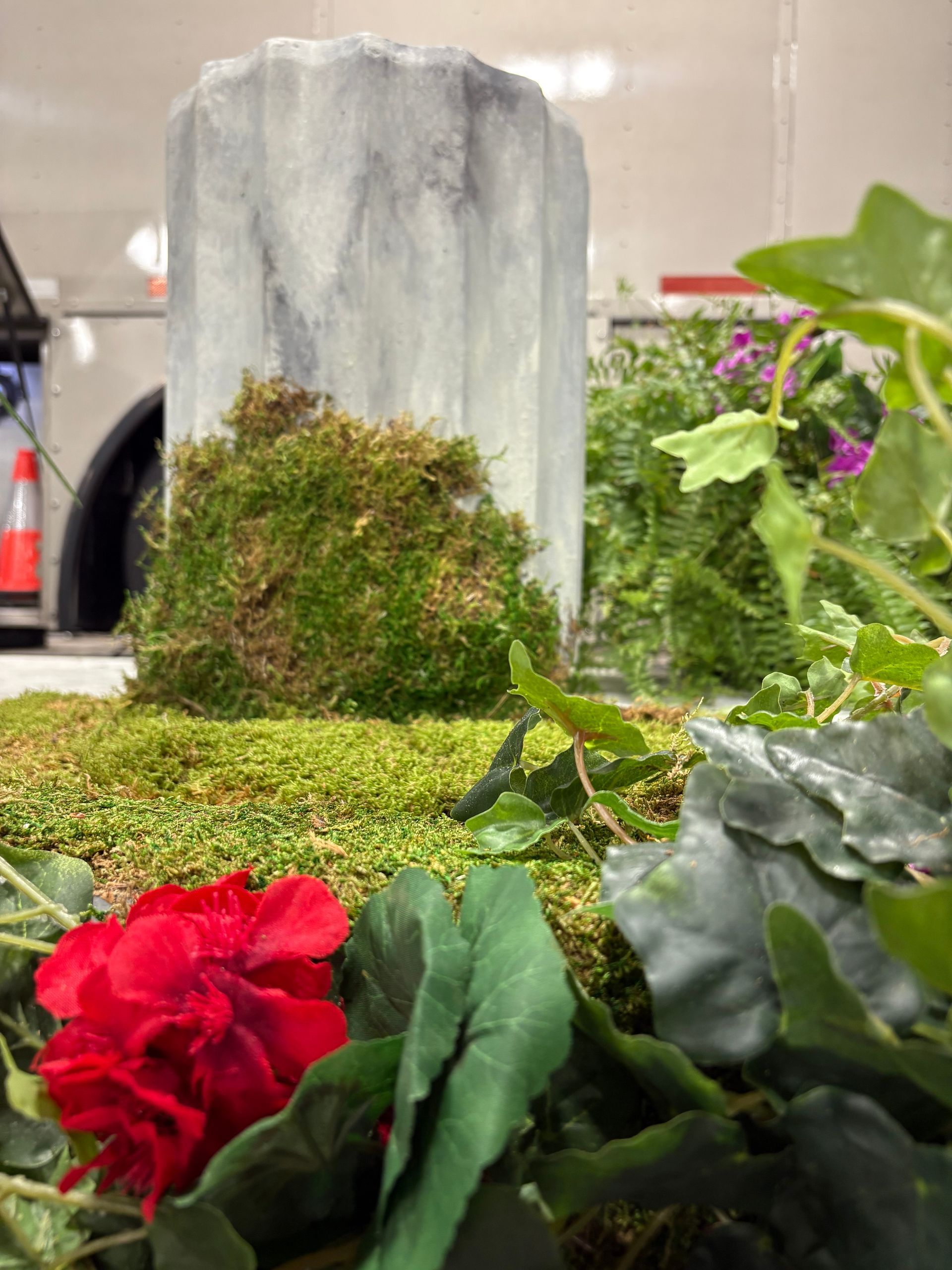 Decorative garden setup with red flower, moss, and a weathered stone monument surrounded by greenery.