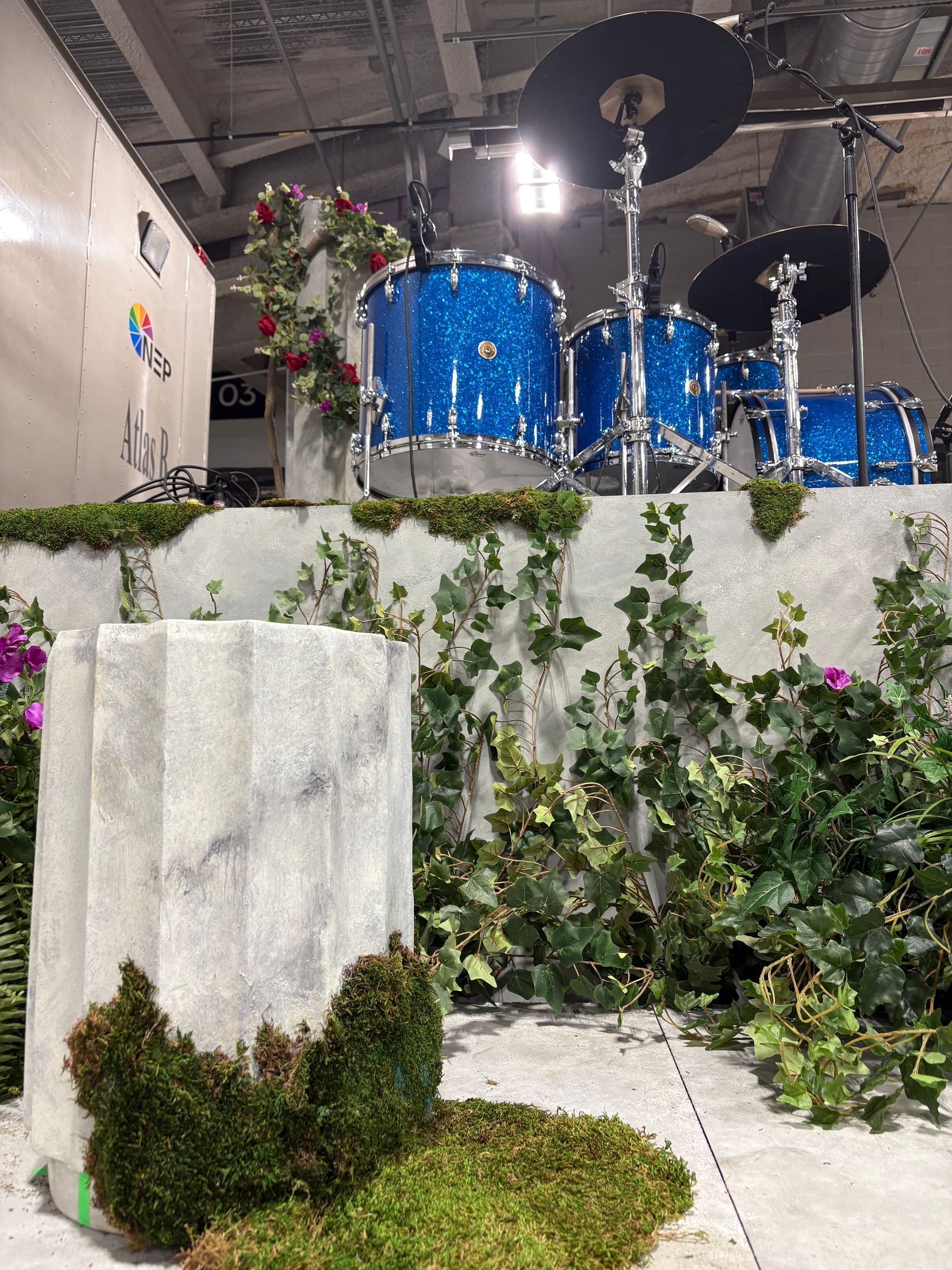 Blue glitter drum kit on a platform surrounded by faux marble columns, moss, ivy, and red and white flowers.