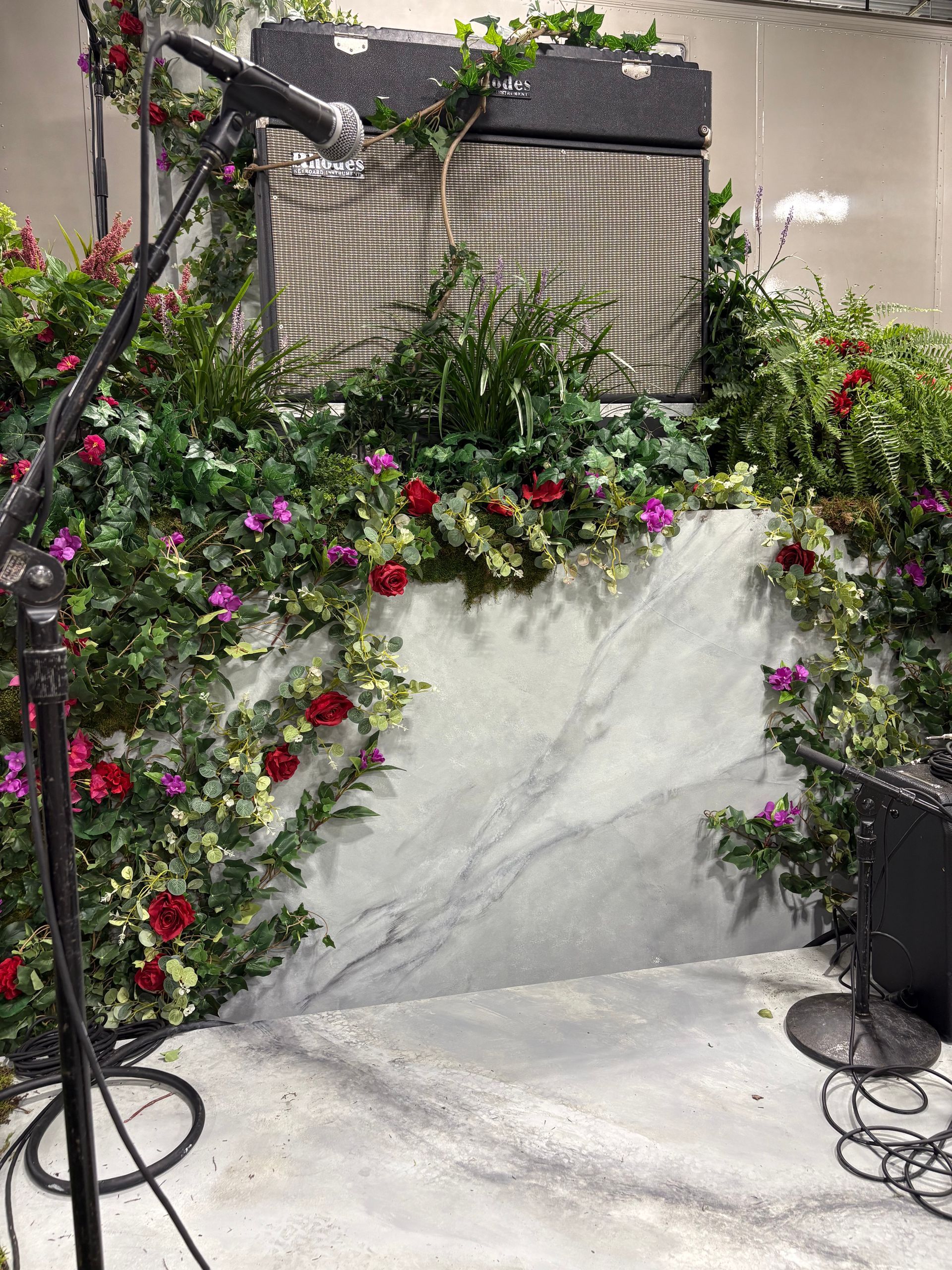 Fender amplifier surrounded by red roses, purple flowers, ivy, and marble slab in a decorative stage setup.