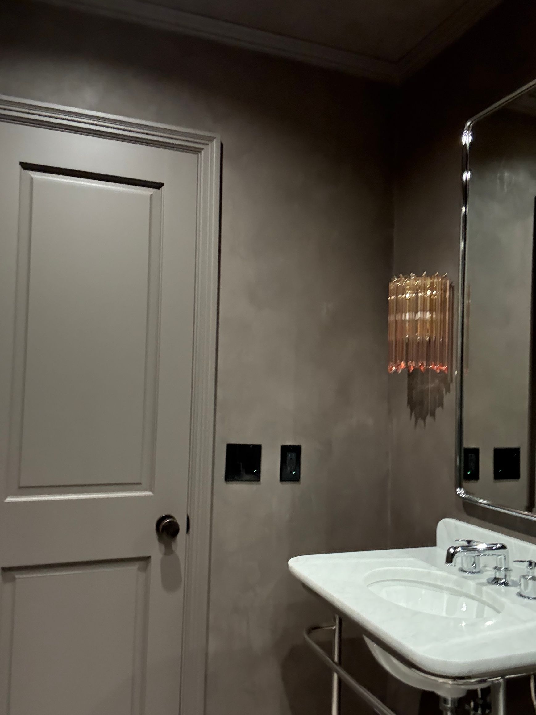 Minimalist bathroom with matte wall, chrome-framed mirror, porcelain sink, and gold vertical light fixture beside black switches.