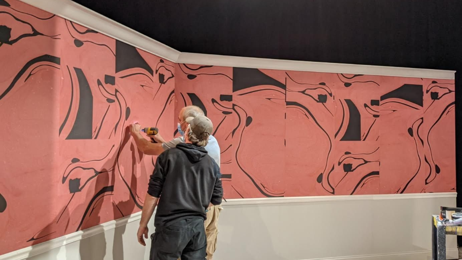 Two people installing abstract red and black wallpaper with swirling anatomical patterns above a white chair rail.