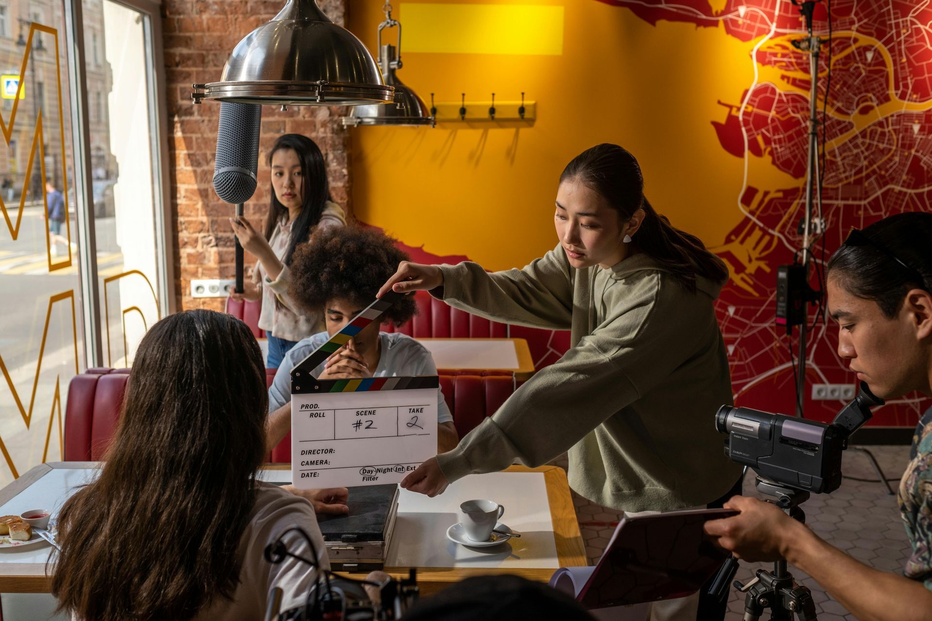Film crew shooting in a cafe; a director holds a clapperboard, others hold camera and boom mic.