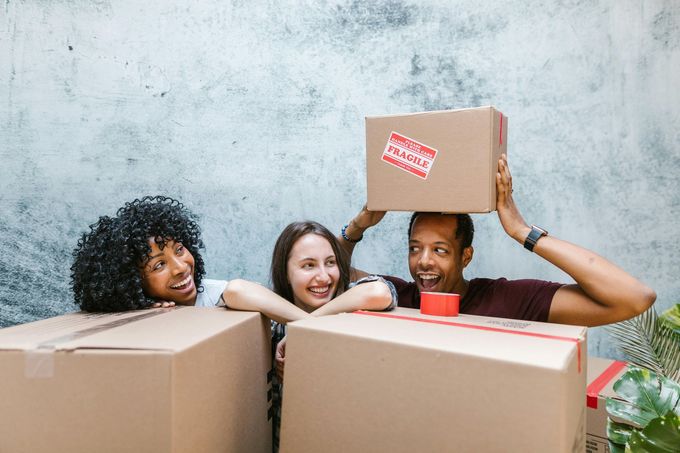 Two women are looking at the man who is holding a fragile box on top of his head in a funny manner.
