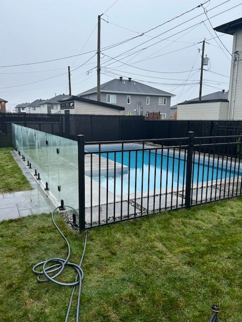 Une clôture noire entoure une piscine. Des sections de verre transparent et de métal noir. Herbe verte et ciel couvert.