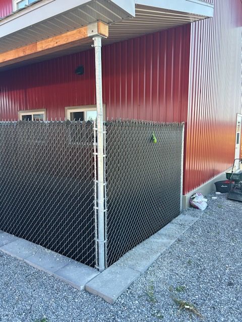 Clôture grillagée noire contre un bâtiment rouge, bordée de gravier gris et de blocs de béton.