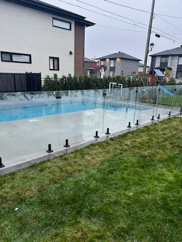 Une clôture en verre entoure une piscine dans un jardin. L'herbe verte et les maisons sont visibles en arrière-plan.