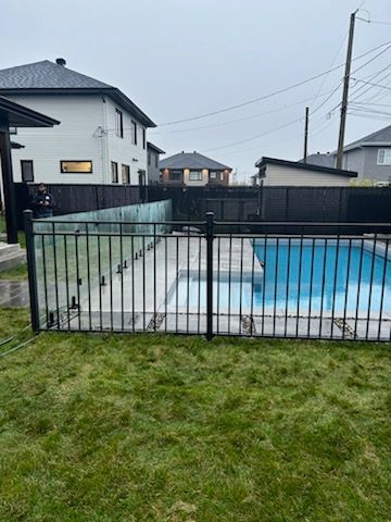 Une piscine avec une clôture en métal noir et des panneaux de verre dans une cour arrière, sous un ciel nuageux.