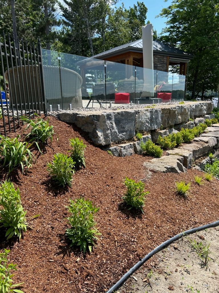 Un espace paysager avec un mur de soutènement, une balustrade en verre et des plantes ; une terrasse avec des chaises rouges est en arrière-plan.