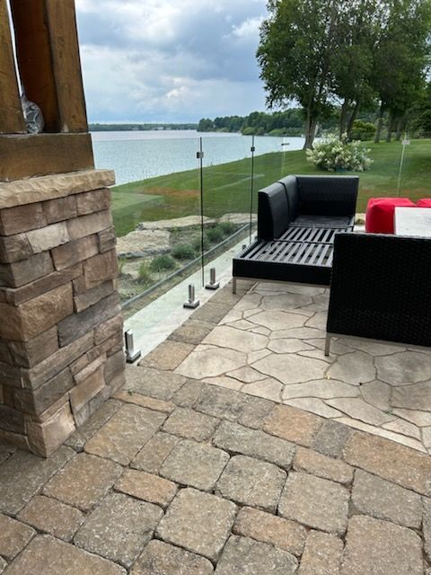 Patio donnant sur un lac avec balustrade en verre, sol en pierre et mobilier d'extérieur noir.