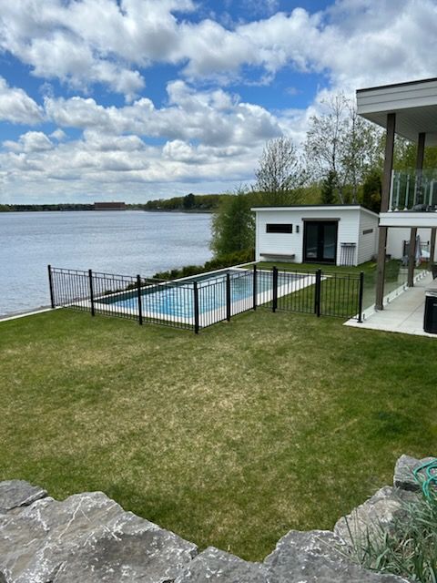 Arrière-cour au bord du lac avec piscine, petit hangar blanc, herbe verte, clôture noire et maison moderne sous un ciel bleu.