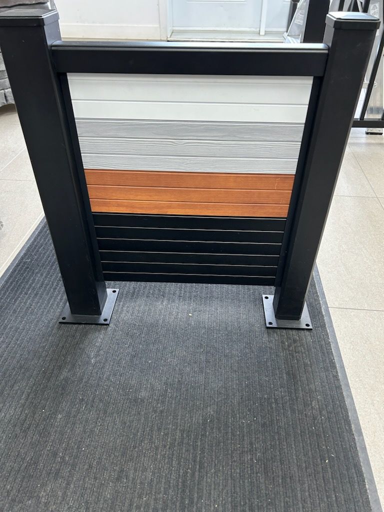 Cadre noir présentant divers échantillons de panneaux de clôture : lattes horizontales blanches, grises, cuivrées et noires, exposées à l'intérieur sur un tapis.