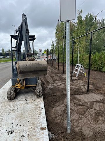 Une excavatrice creusant le long d'un trottoir, avec un panneau de signalisation, une clôture et des arbres en arrière-plan.