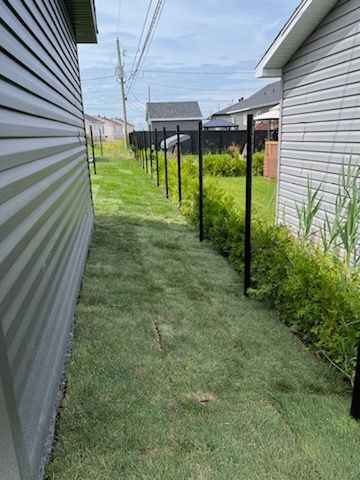 Étroite bande d'herbe entre deux bâtiments, avec des poteaux noirs pour une future clôture.