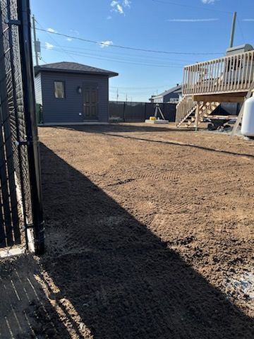 Une cour arrière avec un sol foncé fraîchement labouré, une clôture noire, un hangar et une terrasse en bois sous un ciel bleu.