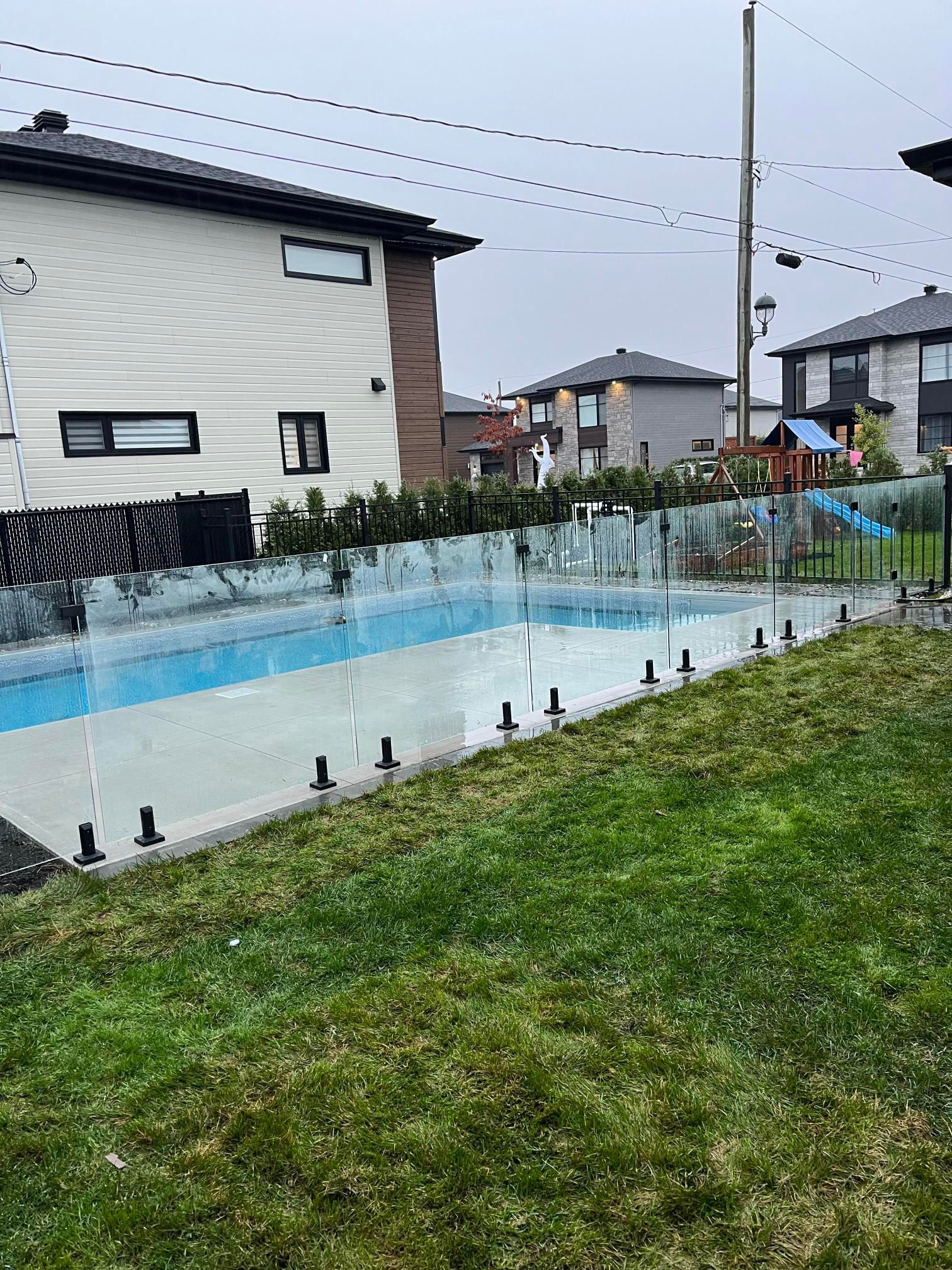 Une cour arrière avec une piscine clôturée en verre, de l'herbe verte et des maisons en arrière-plan par une journée nuageuse.