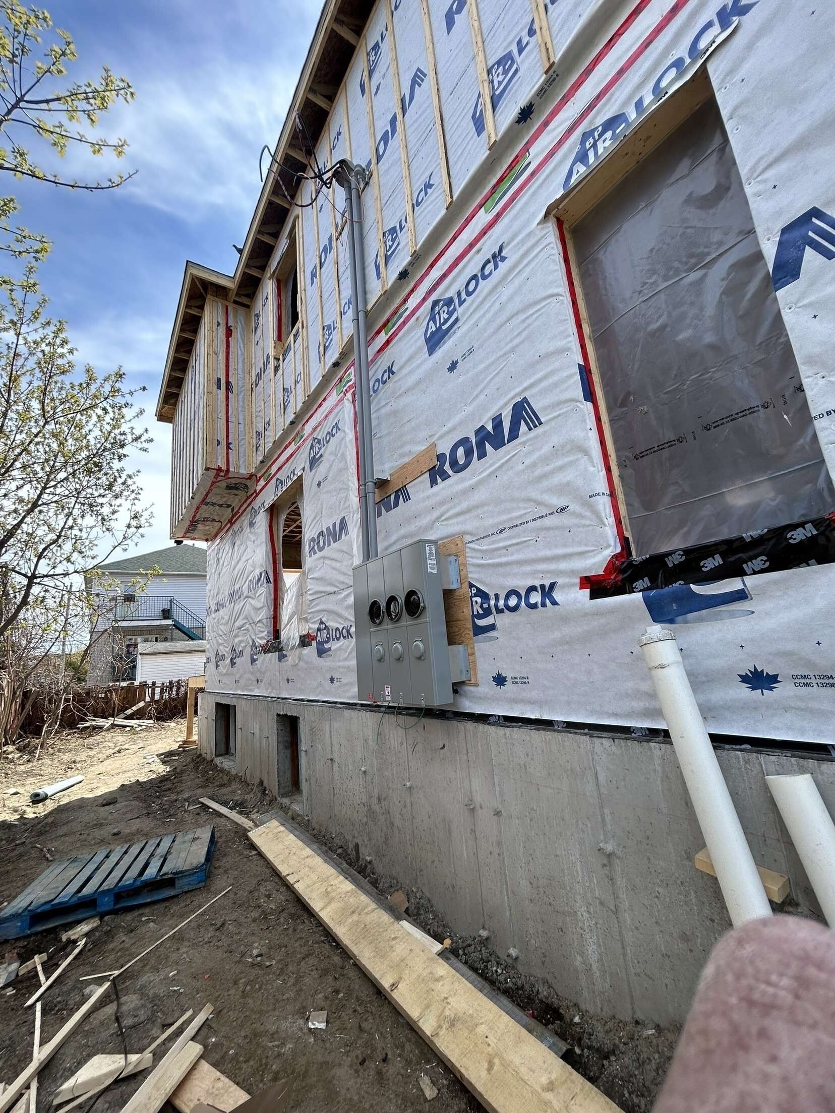 Extérieur d'un bâtiment en construction, comprenant une ossature en bois, une barrière contre les intempéries bleue et des boîtiers de compteurs électriques.