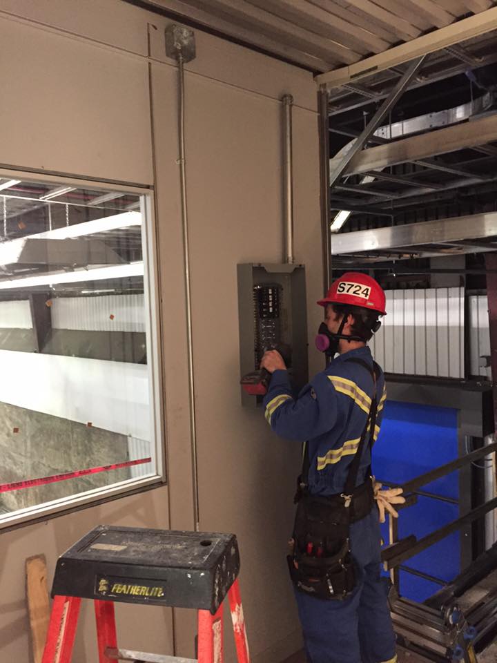 Électricien en combinaison bleue travaillant sur un panneau électrique, portant un casque et un masque respiratoire. À l'intérieur d'un bâtiment en construction.