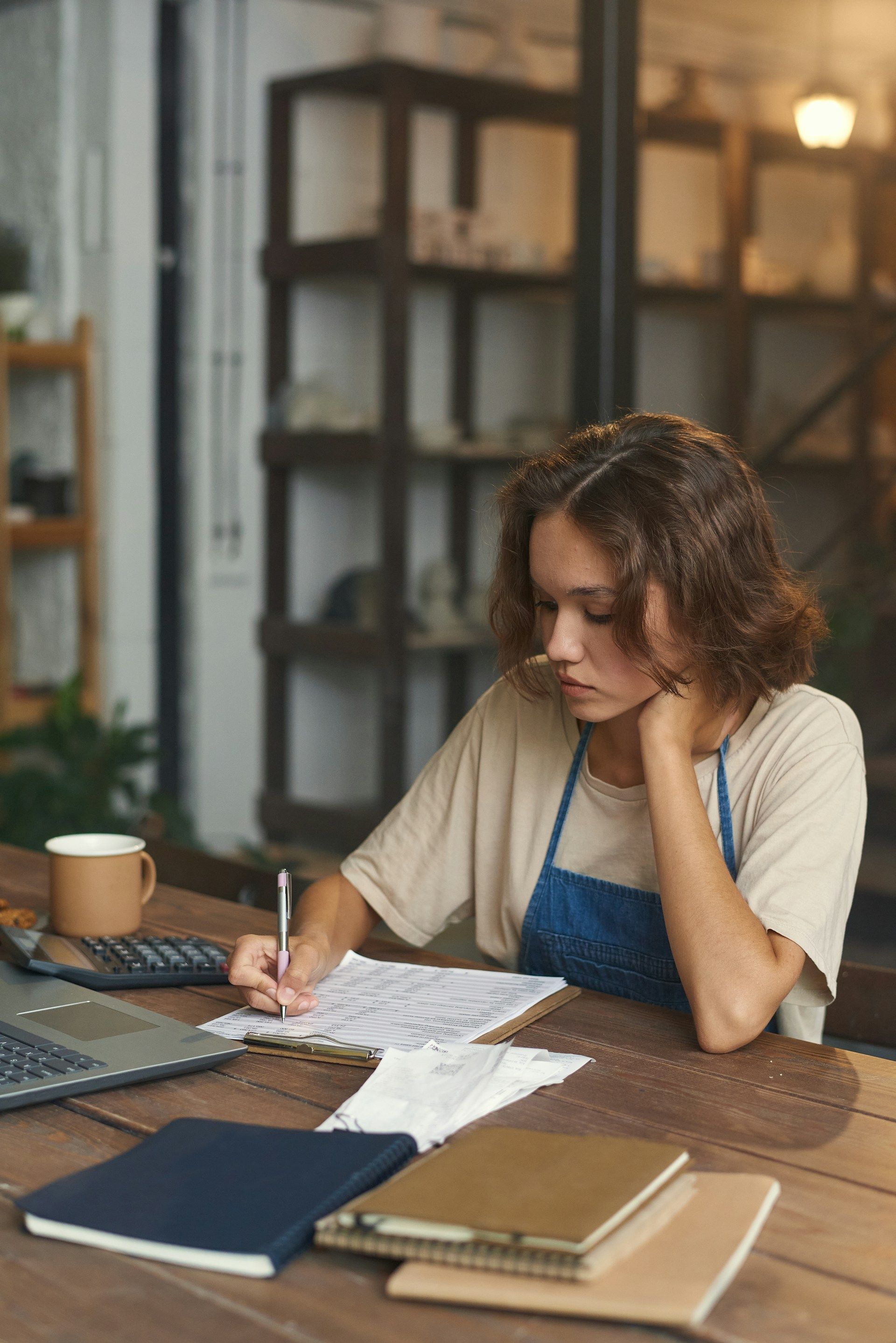 Woman in apron at a table writing, laptop and calculator nearby. Inside a store with shelves in the background.