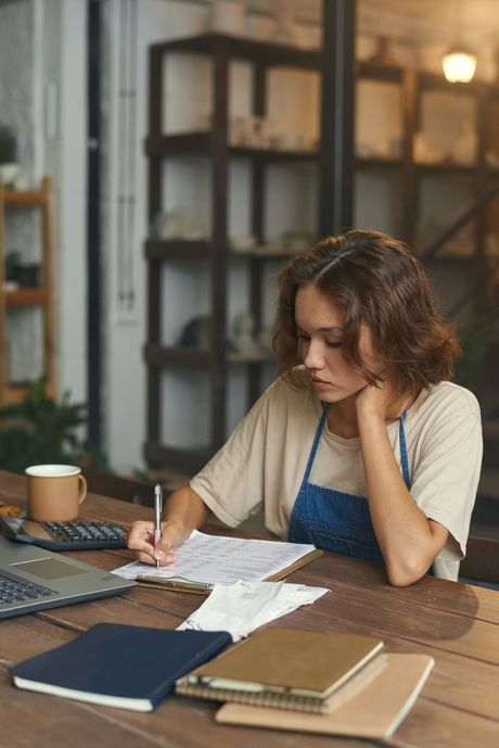 Woman in apron at a table writing, laptop and calculator nearby. Inside a store with shelves in the background.