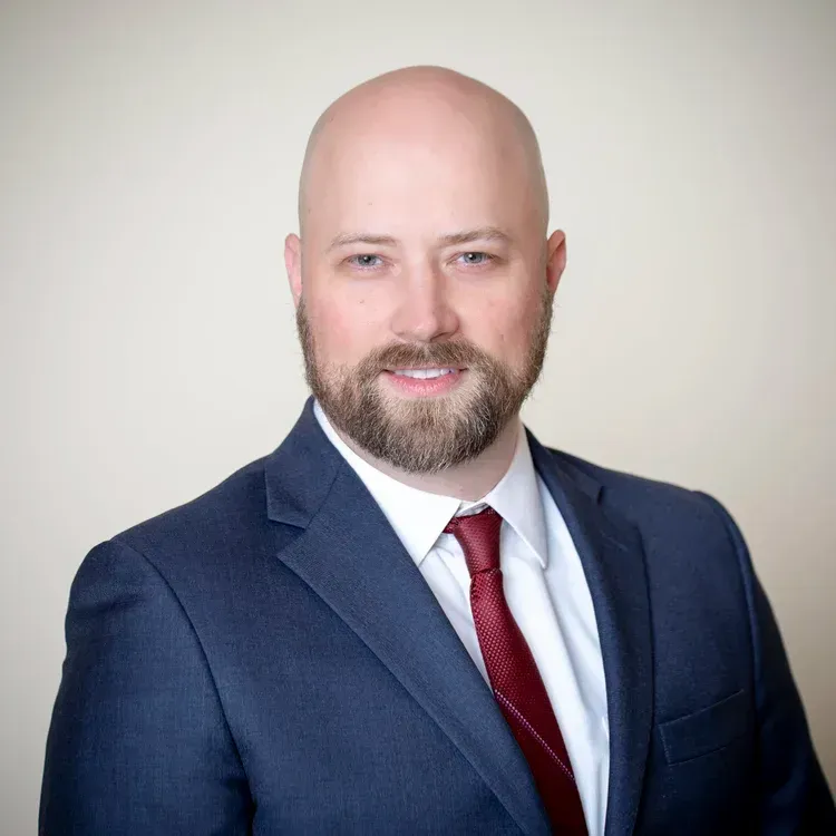 Bald man in blue suit and red tie smiles, posing in front of a neutral background.