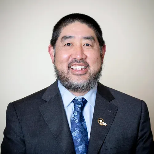 Man in a dark suit smiles, wearing a patterned blue tie and a lapel pin, against a neutral background.