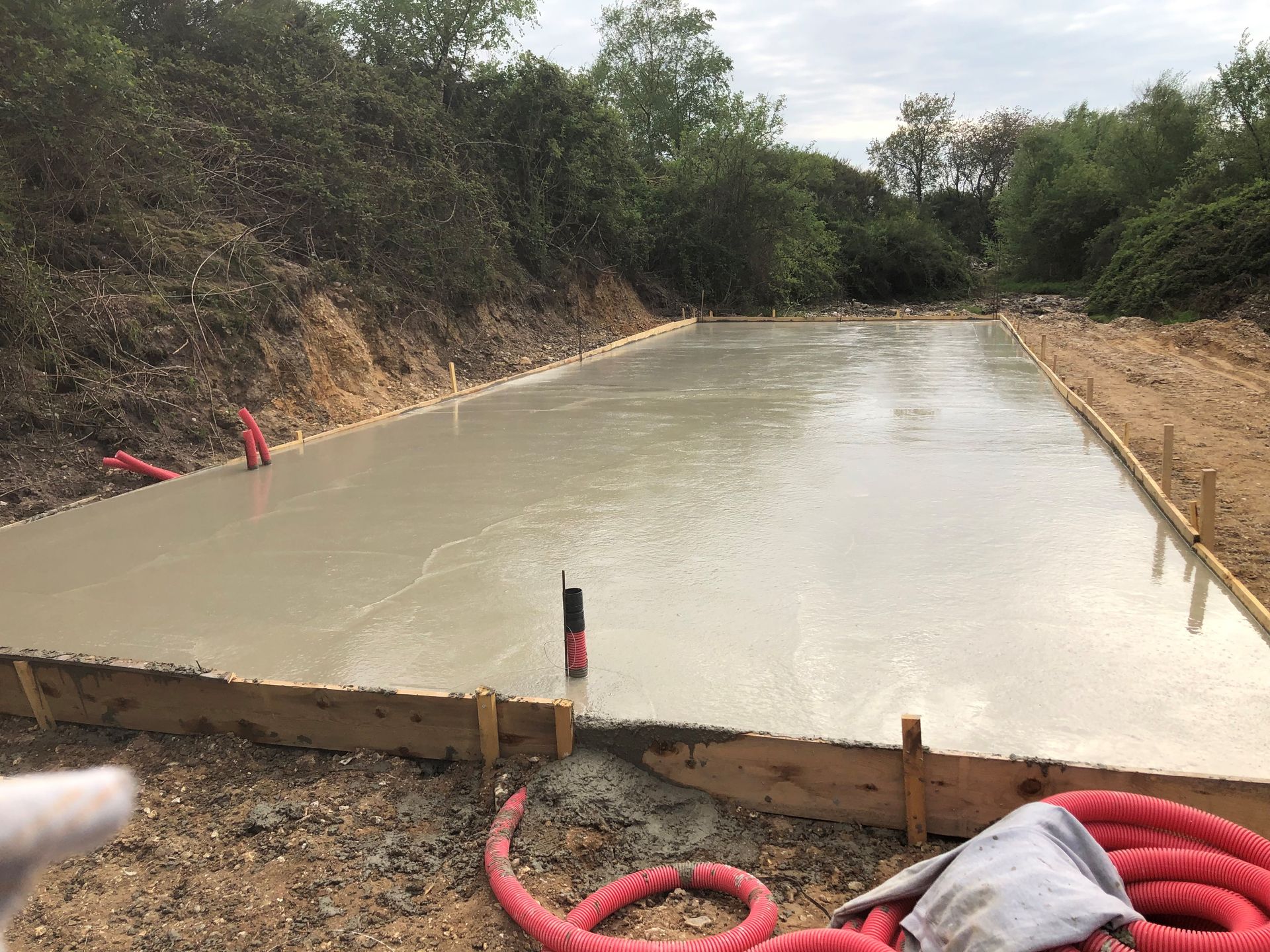 Dalle de béton fraîchement coulée au bord d'un chemin de terre, avec des tuyaux rouges et des piquets dans une zone boisée