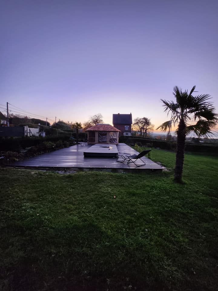 Terrasse extérieure au crépuscule avec une petite structure blanche, une surface mouillée et réfléchissante, un palmier et un premier plan herbeux.