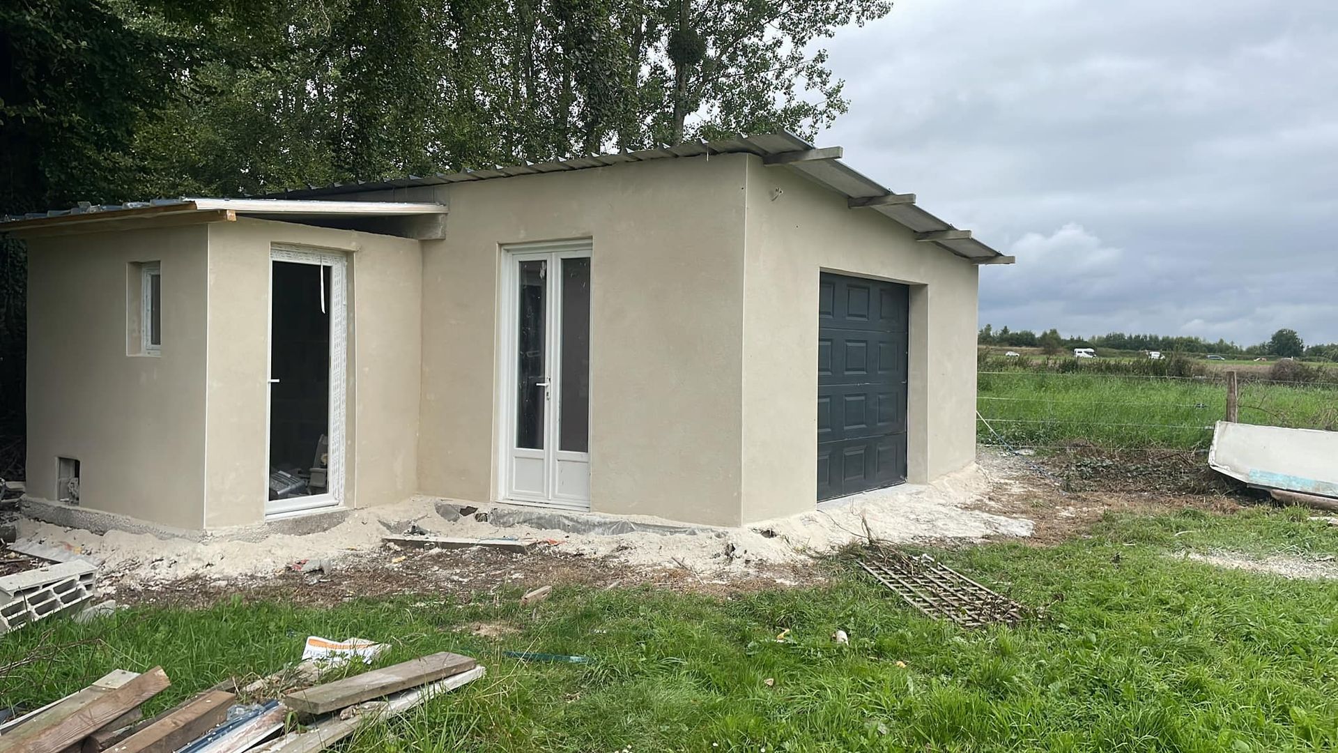 Petite maison beige en construction, portes ouvertes, située dans un champ herbeux à la campagne.
