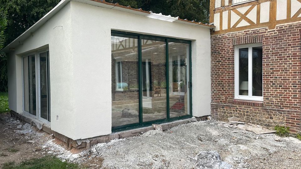 Extension moderne blanche avec de grandes portes vitrées, jouxtant une maison en briques et entourée de gravier.