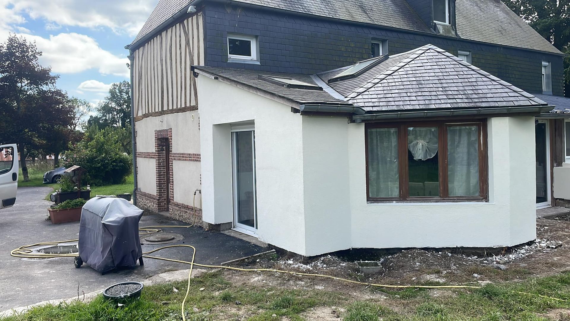 Maison blanche partiellement rénovée avec toit gris et objets couverts dans une allée