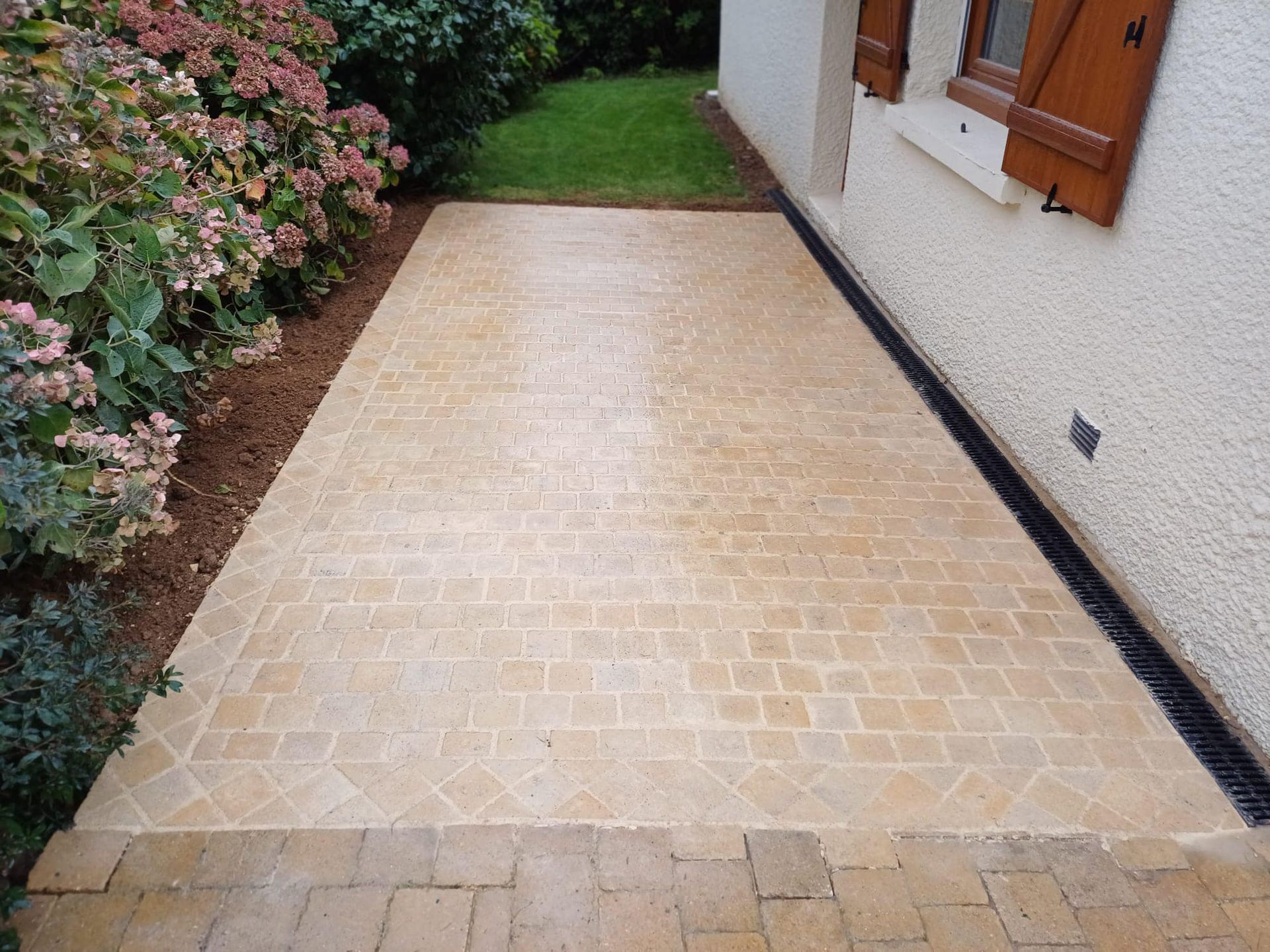 Allée en briques beiges longeant une maison blanche, bordée de haies et de fleurs