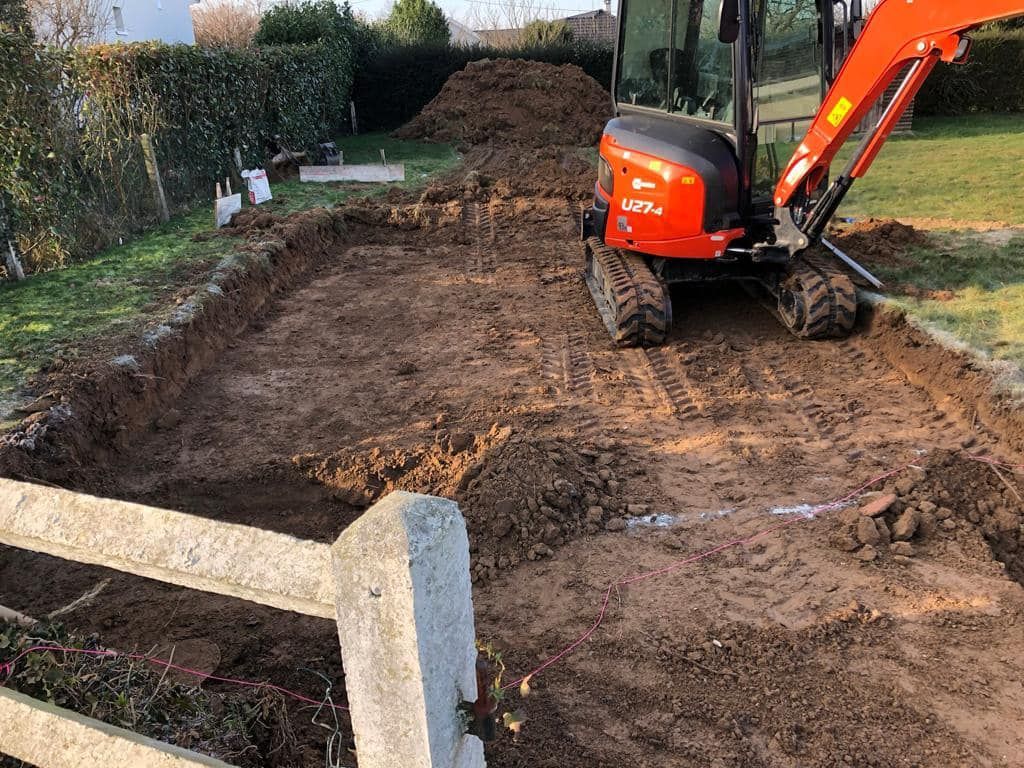 Une pelleteuse orange creuse un jardin boueux, avec un monticule de terre et une haie à proximité.