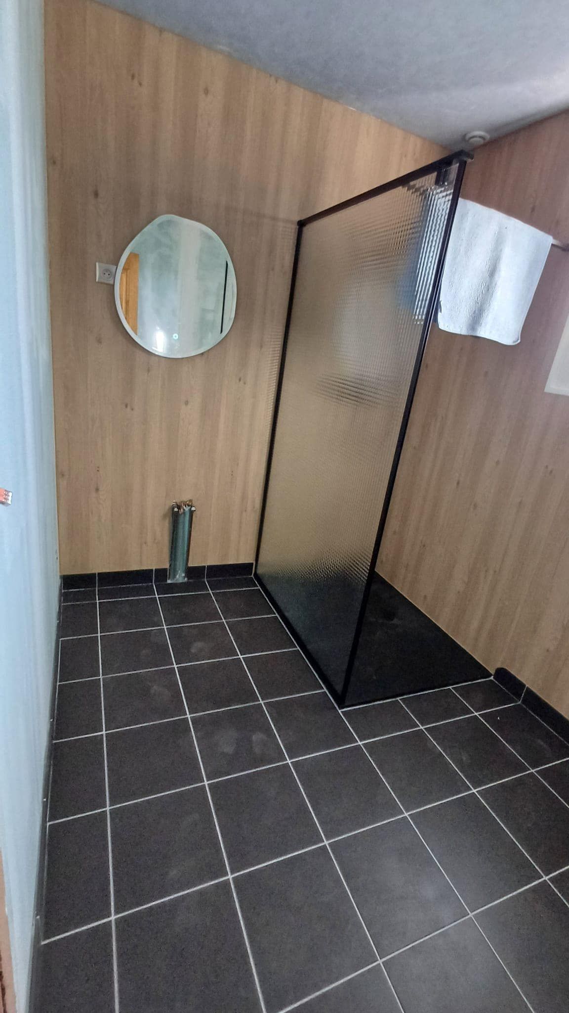 Petite salle de bains avec murs lambrissés, miroir rond, sol carrelé noir et porte de douche en verre avec serviette.