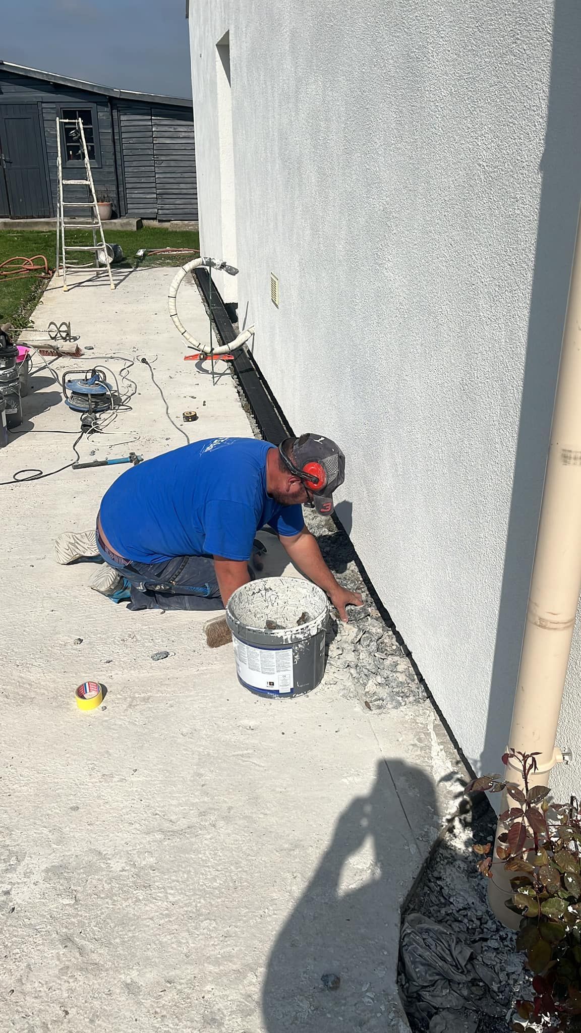 Un ouvrier peint un mur extérieur à côté d'une maison, agenouillé sur du béton avec un seau et des outils.